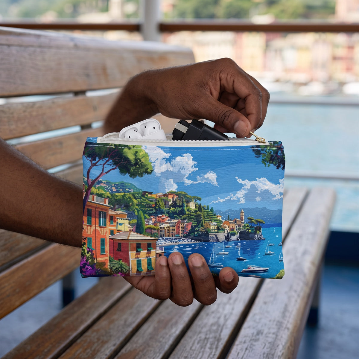 Small Portofino Coastal Escape accessory pouch (8.35 × 5.98 inches) in use: man zips up pouch containing earbuds and a small charger, harbor blurred. Lifestyle mockup with hands showing scale.