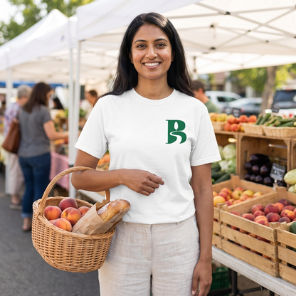 A smiling woman with long dark hair wears the white Bliss Road Studios embroidered tee at a Saturday farmers market, a basket of peaches and bread on her arm