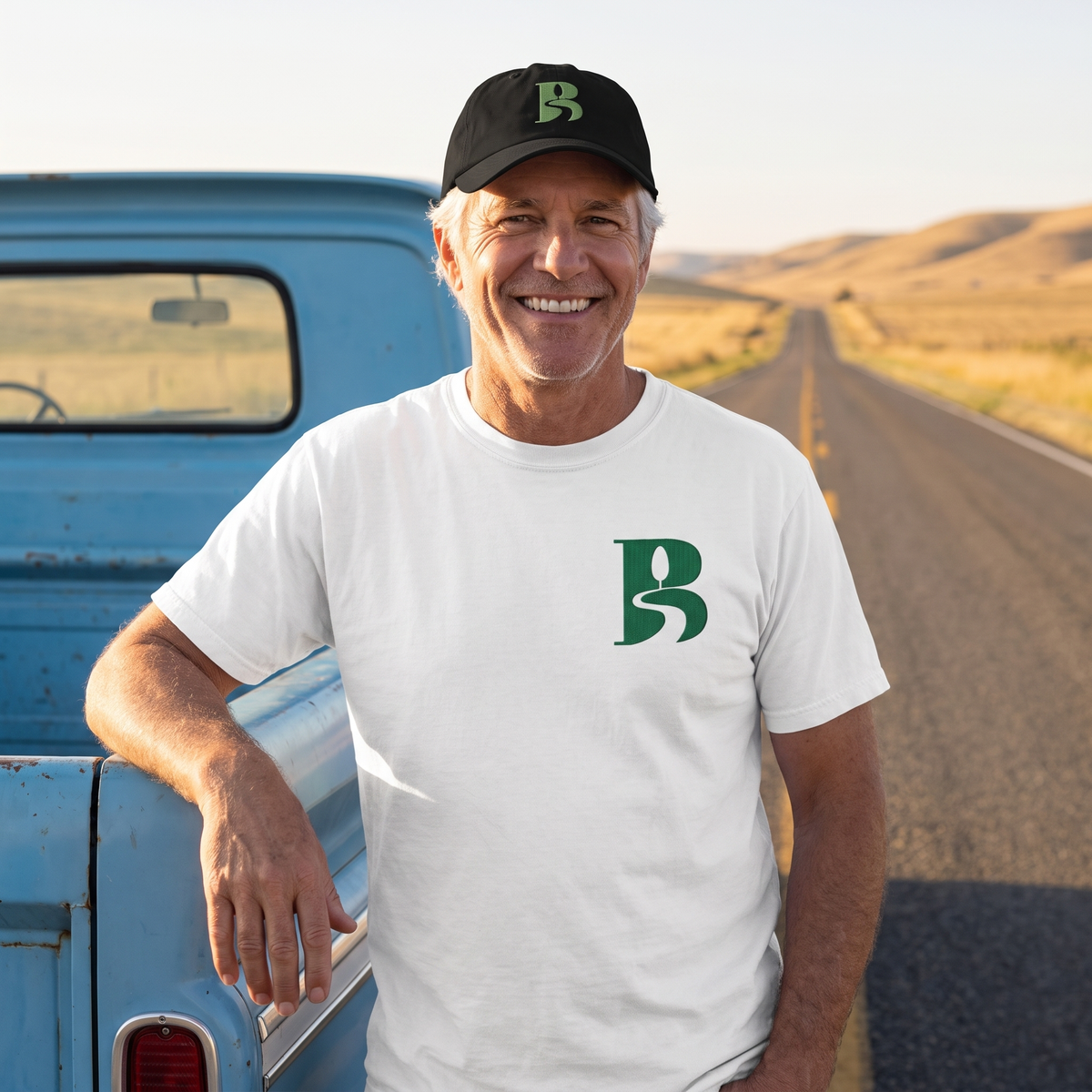 A smiling grey-haired man in his fifties wears the white Bliss Road Studios embroidered tee and black logo cap, leaning on a vintage blue pickup where an open two-lane road runs toward golden hills